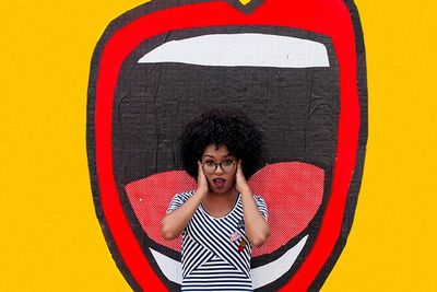 a black woman with glasses covering her ears while standing in front of a mural with a mouth and a yellow background