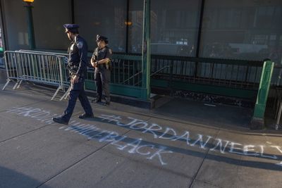 two police officers stand in front of a NYC subway entrance with #JORDANNEELY spray-painted on the ground