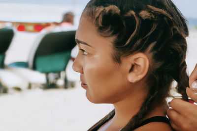 Latin woman braids the hair of a younger companion against the backdrop of a Caribbean beach.