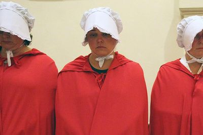 Four women dressed in red robes with white caps covering their faces look down solemnly