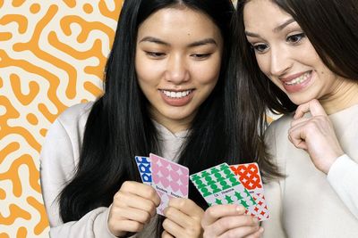 Two Latina women happily playing a board game, holding cards in their hands and enjoying the moment.
