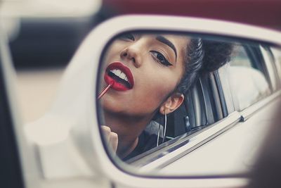 woman with big hoop earrings applying red lipstick on a car rearview mirror