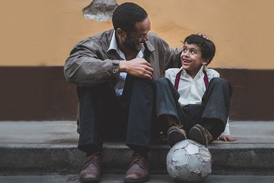 A father and his son sitting on a sidewalk smiling to each other while the boy has a soccer ball under his feet