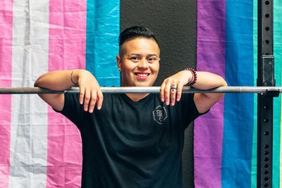 Fitness Coach Stands Facing Camera with hands over bar bell with weights on it in front of a Pride flag